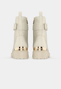 Beige Stiefeletten mit geprägtem Material, goldfarbenen Beschlägen, flacher Sohle und einem Riemendetail am oberen Teil.