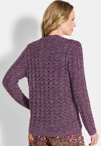 Eine Frau mit welligem blonden Haar trägt einen strukturierten lila Strickpullover und bunt gemusterte Hosen, seitlich vor einem schlichten Hintergrund.