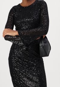 Robe noire à paillettes avec des manches longues, un col rond et une silhouette ajustée. Présente des paillettes brillantes créant une surface texturée. Un sac à main noir est inclus.