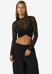 Fekete csipke hosszú ujjú crop top virágmintával, magas nyakkivágással és csavart elülső dizájnnal, laza fekete nadrággal párosítva.