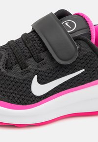 Zwarte sportieve schoen met een bovenwerk van mesh, voorzien van een klittenbandsluiting, witte Nike swoosh en roze accenten op de zool. Geprofileerde stof zorgt voor grip.
