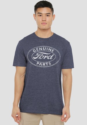 Mann trägt ein dunkelblaues T-Shirt mit weißem "Genuine Ford Parts"-Logo, steht vor schlichtem hellgrauem Hintergrund.