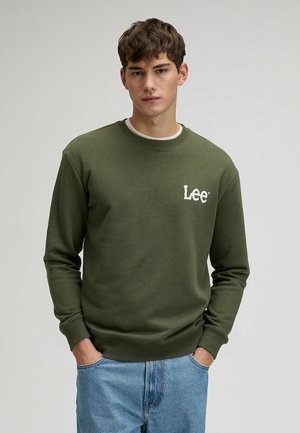 Młody mężczyzna z krótkimi brązowymi włosami, ubrany w oliwkową bluzę Lee i jasnoniebieskie dżinsy, stoi ze złożonymi rękami w kieszeniach.