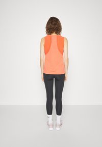 Persoon met golvend bruin haar staat met de rug naar de camera en draagt een mouwloos oranje sporttop, zwarte legging, witte Nike sokken en roze sneakers.