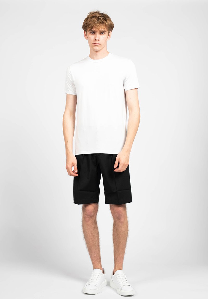 T-shirt bianca in cotone, pantaloni corti neri con un design strutturato e sneakers bianche. Il modello si trova di fronte a uno sfondo chiaro e uniforme.
