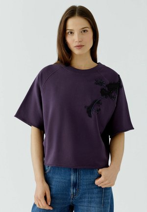 Jonge vrouw met stijl bruin haar draagt een losse donkerpaarse top met zwarte bloemenborduur en blauwe spijkerbroek tegen een effen lichte achtergrond.