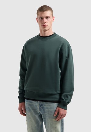 Jonge man met kort haar die een donkergroene sweatshirt over een zwart shirt en lichtblauwe versleten jeans draagt, staande tegen een effen achtergrond.