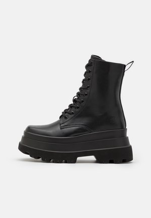 Botte de combat montante en cuir noir avec semelle plateforme épaisse, lacets à l'avant et languette à l'arrière sur fond blanc.