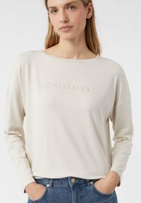 Beige tričko s dlouhým rukávem a volným střihem, na kterém je decentně vytištěn nápis "MINIMALIST" přes hrudník. Viditelné modré džíny.