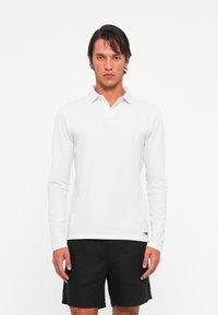 Man standing facing forward wearing a white long-sleeve polo shirt and black shorts against a plain white background.