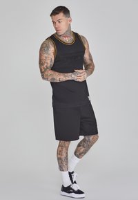 Svart ärmlös tank top med guldkant och logotyp, matchad med svarta shorts. Innehåller tatueringar på armar och ben.