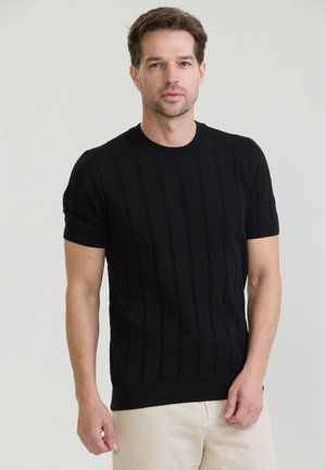 Mand med kort brunt hår iført en sort kortærmet striktrøje og lysebeige bukser mod en ensfarvet lys baggrund.