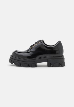 Chaussure noire brillante à lacets avec semelle épaisse à crampons et œillets dorés, vue de profil sur fond clair uni.