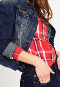 Veste en denim avec deux poches poitrine, portée par-dessus une chemise à carreaux rouge. Jean foncé avec des détails de couture, mettant en avant une superposition décontractée.