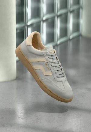 Hellgrauer Wildledersneaker mit beigen Akzenten und Gummisohle, schwebt über poliertem Betonboden in der Nähe einer Glassteinmauer.