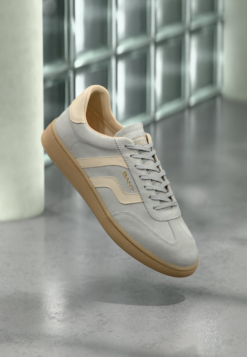 Απαλό γκρι σνίκερ από σουέντ με μπεζ λεπτομέρειες και καφέ σόλα, αιωρείται πάνω από γυαλισμένο τσιμεντένιο πάτωμα κοντά σε τοίχο από γυάλινα τούβλα.