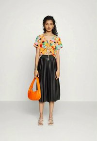 Top cropped floral colorido com laço na frente, saia midi preta plissada e uma bolsa laranja de ombro. Modelo usa sandálias brancas de tiras.