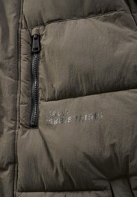 Puffig olivgrön jacka med en svart dragkedja, med texten "HALO 1968/19721981" tryckt i grått på tyget.