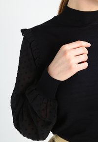 Mujer vistiendo una blusa negra con mangas abullonadas y transparentes de lunares, y con un borde rizado, levantando la mano cerca de la clavícula contra un fondo blanco.