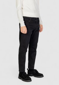 Pantaloni in denim neri con vestibilità slim, dotati di zip e design a cinque tasche. Abbinati a una maglietta bianca a maniche lunghe e scarpe nere.