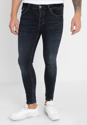 Mann trägt dunkle enge Jeans und weiße Turnschuhe, steht mit entspannt an den Seiten hängenden Händen vor einem schlichten Hintergrund.