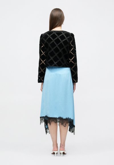 Μαύρο βελούδινο σακάκι με ραμμένη επένδυση και φουσκωτά μανίκια, σε συνδυασμό με φούστα ανοιχτού γαλάζιου χρώματος με δαντελένιο τελείωμα, φορεμένα με λευκά παπούτσια με τακούνι.