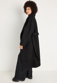 Cappotto di lana nero e morbido con colletto a scialle, maniche lunghe e vita con cintura. Presenta una superficie testurizzata e un design di lunghezza media.