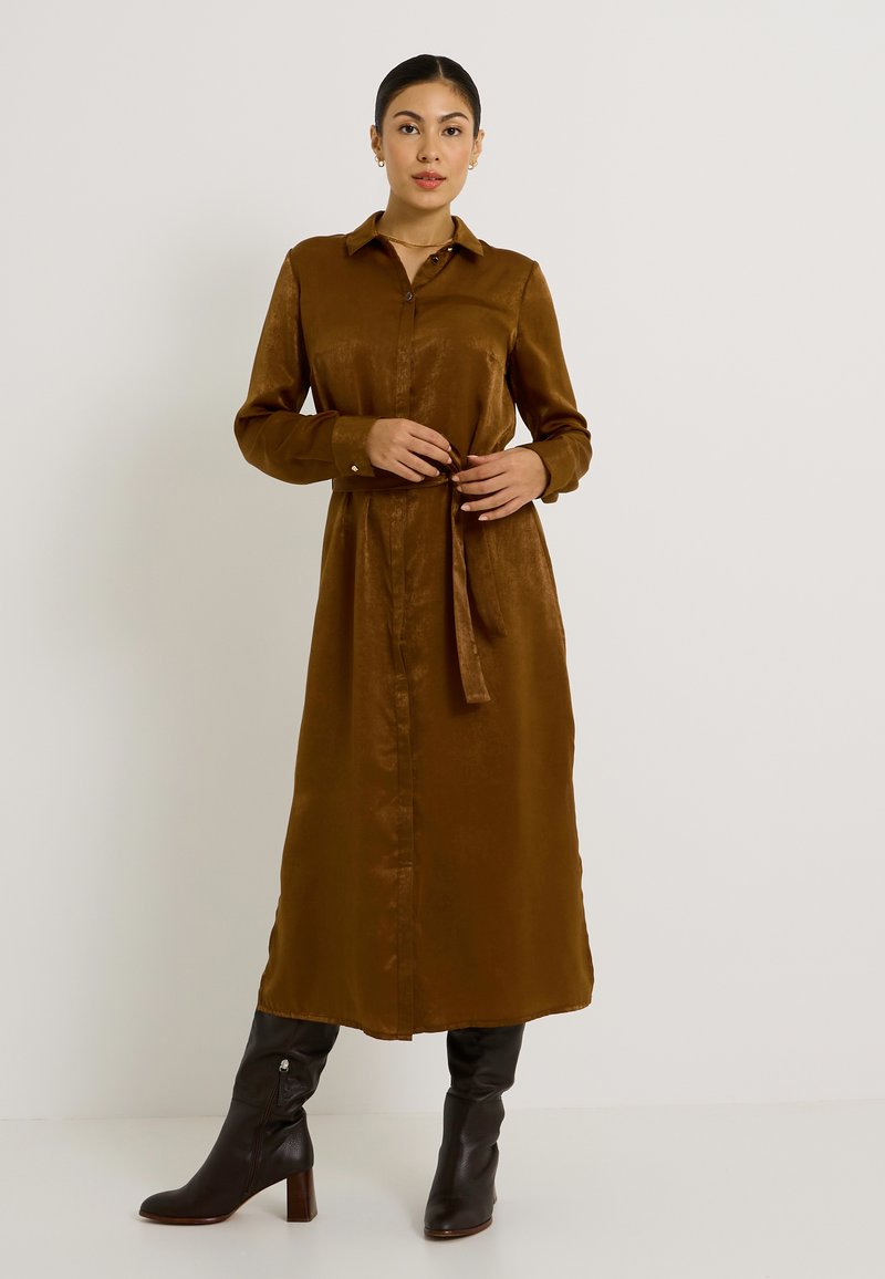 Vestido de camisa de manga larga en bronce, de tejido suave, con botones en la parte delantera, cuello y cintura ajustable, combinado con botas negras de tacón altas hasta la rodilla.