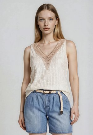 Frau mit langen blonden Haaren, die ein ärmelloses cremefarbenes, strukturiertes Top mit Spitzen-V-Ausschnitt und hellblaue Jeansshorts mit geflochtenem Gürtel trägt.