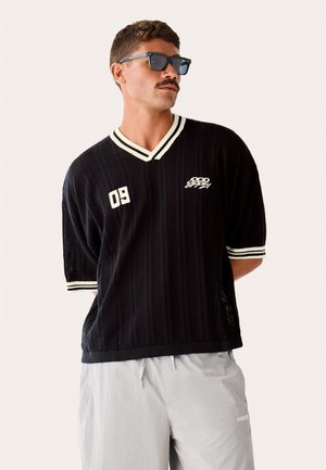 Man with mustache wearing black knit V-neck shirt with white stripes and "09" and logo, paired with light gray pants and sunglasses.