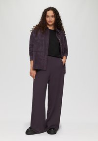 Langere mouwen, donkerpaarse corduroy shirt met een voorzak over een zwart t-shirt, gecombineerd met loszittende, donkerpaarse broek en zwarte schoenen.