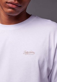 Witte katoenen T-shirt met een ronde halslijn en korte mouwen. Logo "Dolly Noire" in lichtbruin geborduurd op de borst.