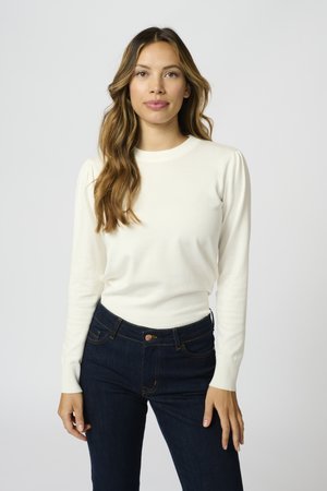 Cremefarbener Pullover mit langen Ärmeln, geripptem Ausschnitt und Bündchen, kombiniert mit dunkelblauen Denim-Jeans, die orangefarbene Nähte und einen vorderen Knopf haben.