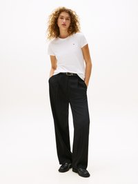 White t-shirt with a small logo, black wide-leg trousers, and black shoes. Model has curly hair and hands in pockets. Simple, monochrome look.