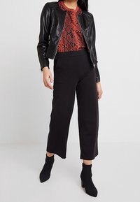 Veste courte en cuir noir portée sur un haut rouge à imprimé serpent, associée à un pantalon noir à jambes larges et des bottines noires pointues. Textures lisses.