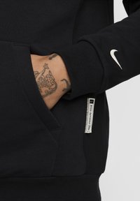 Felpa nera con tasca a marsupio, polsini a coste e logo Nike bianco sulla manica. Il tessuto sembra morbido e testurizzato.