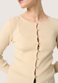 Ribbad beige cardigan med rund halsringning och silverknappdetaljer fram, med en vågig knappslå och snug passform.