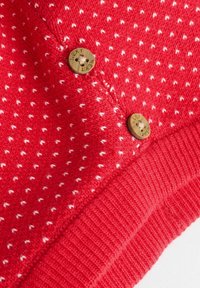 Rotes gestricktes Kleidungsstück mit einem weißen gepunkteten Muster und einem gerippten Saum. Verfügt über zwei runde Holzknöpfe mit eingraviertem Text.