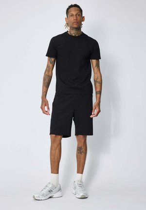 Homme avec des tatouages portant un t-shirt noir et un short, des chaussettes blanches et des baskets argentées, debout devant un fond blanc uni.