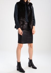 Gilet in pelliccia sintetica nera sopra un top blu verde scuro, abbinato a una pencil skirt in pelle nera e stivaletti neri con tacco a blocco.