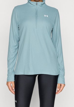 Donna che indossa un top azzurro a manica lunga con zip al quarto e leggings neri con loghi Under Armour su entrambi i capi.