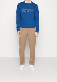 Blå sweatshirt med "BOSS"-logotyp, beige byxor och vita sneakers. Slät tyg, avslappnad passform, avslappnad design. Lagerkrage detalj synlig.