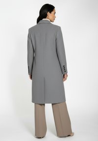 Grå, struktureret lang frakke med klapslag, to rygslids og mørke knapper. Sammen med beige bukser med vidde ben.