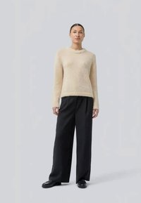 Un pull en tricot de couleur crème avec un col rond, porté avec un pantalon large noir et des chaussures noires, sur un fond clair et uni.