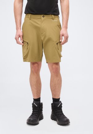 Férfi, aki világosbarna cargo rövidnadrágot visel cipzáras zsebekkel, fekete zoknival és fekete magas szárú csizmával, egyszerű háttér előtt áll.