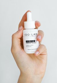 Biała szklana buteleczka z pipetą i białą nakrętką, oznaczona etykietą "IDO LAB TROXACALM", zawierająca serum do kontroli czerwienienia się na twarz, trzymana w dłoni na jasnym tle.