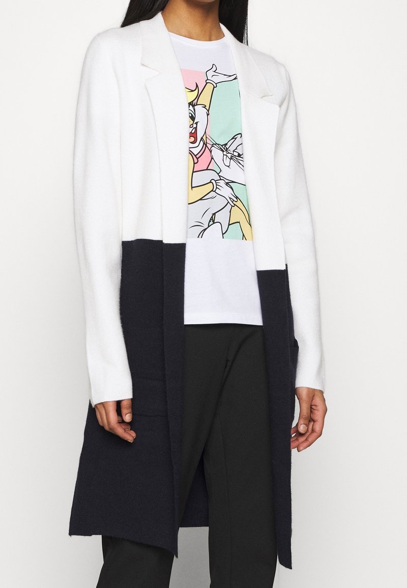 Cardigan lungo in bianco e nero con colletto a revers, tessuto testurizzato, apertura frontale e tasche laterali, indossato sopra una t-shirt grafica.