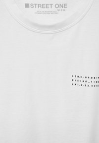 Vit bomull t-shirt med rund halsringning och svart tryckt text på vänster bröst, inklusive koordinater och design detaljer.