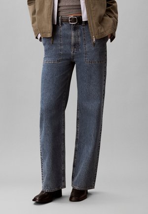 Blå højtaljede vidde jeans med forlommer båret med bælte, brune støvler, en brun top og en tanjakke.