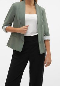 Zielony żakiet o długości rękawa do połowy łokcia, z mankietem w paski, zestawiony z białą bluzką i czarnymi spodniami. Miękka faktura i dopasowany krój.